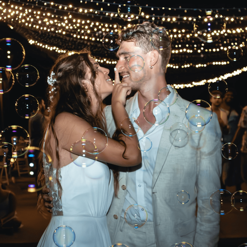 Bride and groom kiss in front of string lights and floating bubbles
